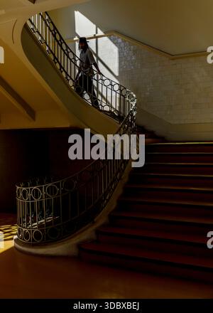 Eine Person läuft eine geschwungene Treppe hinunter mit einem Metallgeländer. Die Treppe ist in einem Gebäude mit gekachelten Wänden und einem gelben Boden. Stockfoto