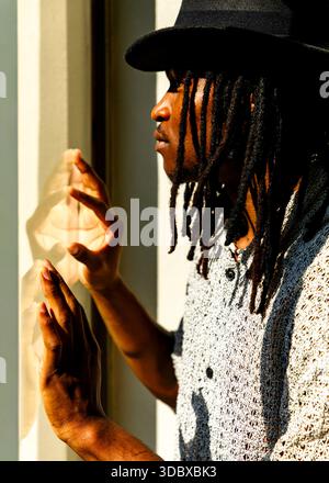 Ein Mann mit schwarzem Hut und gemustertem Hemd berührt ein Fenster mit den Händen. Stockfoto