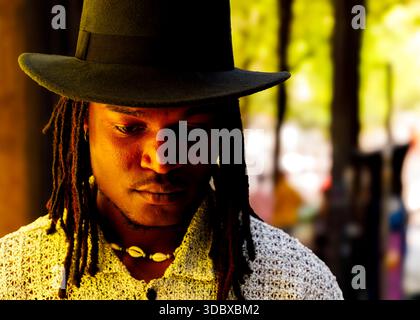 Ein Mann mit Dreadlocks, der einen schwarzen Hut und ein weißes Hemd trägt und nach unten blickt. Stockfoto