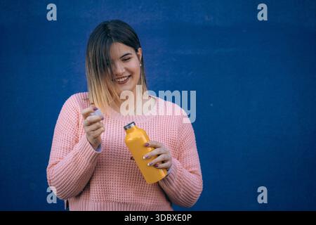 Lächelnde junge Frau in rosa Pullover, die eine gelbe wiederverwendbare Wasserflasche öffnet und vor einer leuchtend blauen Wand steht. Stockfoto