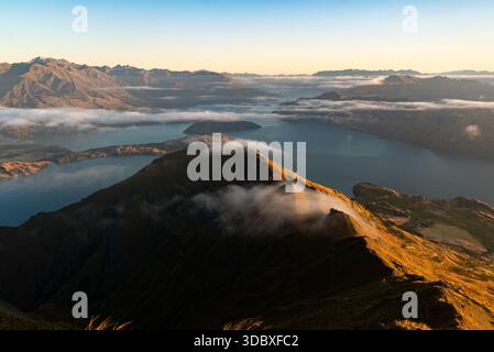 Blick auf einen majestätischen Berggipfel, der sich über einem Wolkenmeer erhebt, mit einem See, der sich zu weit entfernten Gipfeln unter weichem Himmel erstreckt, Roys Peak, Otago Region, Stockfoto