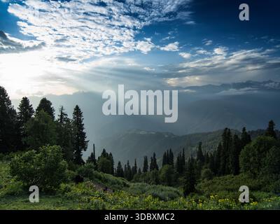 Blick auf Sonnenstrahlen, die durch dramatische Wolken über eine grüne Berglandschaft mit Bäumen durchdringen, Sri Paye, Khyber Pakhtunkhwa, Pakistan. Stockfoto