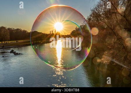 Magischer Sonnenuntergang in der Seifenblase, die tiefstehende Wintersonne taucht die Isar in ein goldenes Licht, München, 18. Dezember 2025 Deutschland, München, 18. Dezember. Dezember 2025, Sonnenuntergang in der Seifenblase, wie durch eine gläserne Christbaumkugel strahlt die tiefstehende Wintersonne und taucht die Isar in einem magischen Licht, ein strahlender Wintertag geht zu Ende, das Heizkraftwerk Süd am Horizont, ein Mann auf der Reichenbachbrücke lässt die Seifenblasen zur Freude der Passanten in den blauen Himmel steigen, Donnerstagnachmittag, Temperaturen um die 10 Grad, Weihnachtszeit, Advent Stockfoto