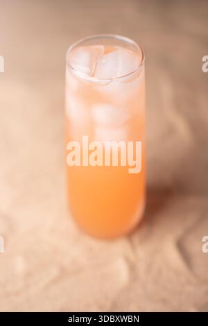 Nahaufnahme eines erfrischenden orangetönten Getränks in einem klaren Glas mit Eiswürfeln, das vor einem leicht verschwommenen hellbeigen Hintergrund steht und eine Stockfoto