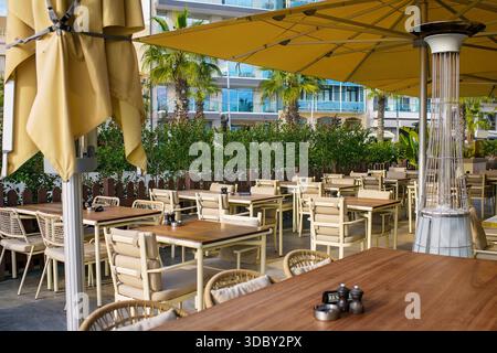 Außenterrasse des Restaurants mit reservierten Holztischen, Korbsesseln, Sonnenschirmen und Gasheizung vor Palmen und Gebäude Stockfoto