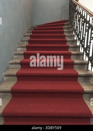 Roter Teppich auf Marmortreppen mit einem schmiedeeisernen Zaun und hölzernem Handlauf auf der rechten Seite und einer weißen Wand auf der linken Seite Stockfoto