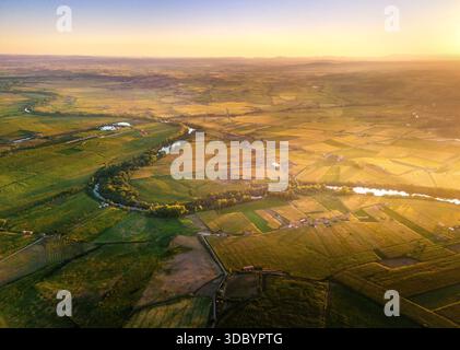 Ein atemberaubender Blick aus der Luft auf einen sich windenden Fluss, der bei Sonnenuntergang durch weite grüne und goldene Felder fließt. Von einer Drohne erfasst, offenbart die Landschaft den bea Stockfoto
