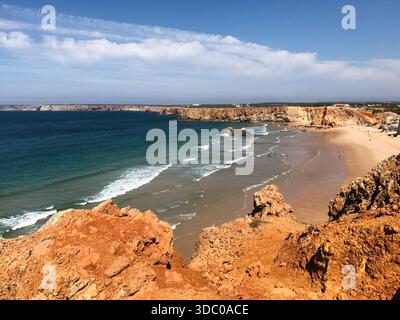 Eine Küstenlandschaft im Süden Portugals zeigt einen breiten Sandstrand, türkisfarbene Atlantikwellen und zerklüftete orangene Klippen unter einem klaren blauen Himmel. Stockfoto