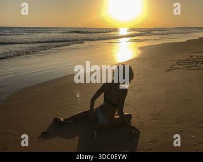 Die Silhouette einer nicht erkennbaren Frau sitzt auf dem Sand vor dem Meer, während die niedrige Sonne hell über dem Wasser und der nassen Küste reflektiert. Stockfoto