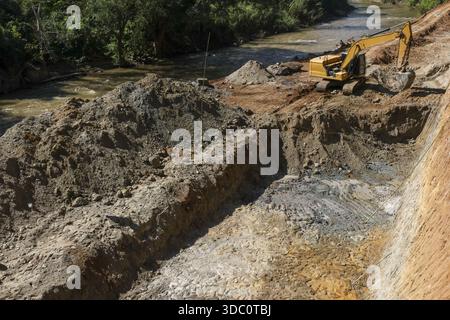 Aktiver gelber Baggerbagger, der ein großes Loch neben dem Flussufer für Baustellen mit Schlamm und Sand grabt und produktive industrielle Energie erzeugt Stockfoto