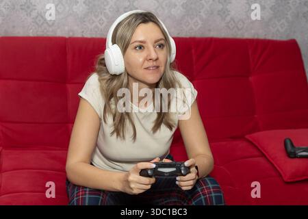 Eine junge Frau sitzt auf einer roten Couch, trägt Pyjamas und weiße Kopfhörer. Sie hält ein Gamepad und konzentriert sich auf ein Videospiel in ihrem Wohnzimmer. Stockfoto