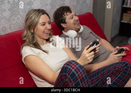 Ein fröhlicher Mann und eine fröhliche Frau sitzen und lachen auf einer roten Couch, während sie mit Handheld-Controllern ein Videospiel spielen. Das Paar trägt einen Pyjama in der gemütlichen Umgebung ihres Zuhauses. Stockfoto