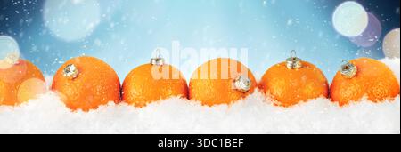 Mandarinen oder Mandarinen in Form von weihnachtstannenschmuck in Schneewehen mit fallendem Schnee und Bokeh auf blauem Hintergrund. Banner Weihnachten im Hintergrund Stockfoto