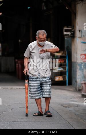 Bangkok, Thailand, 19. November 2025, ein älterer Mann steht in einer schmalen Straße von Talat Phlu und überprüft seine Uhr, während er einen Schirm hält. Der Trubel Stockfoto