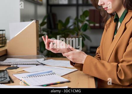 Kaukasische junge Erwachsene Frau, die am Schreibtisch sitzt und das Handgelenk massiert, während sie mit Dokumenten und Notebook arbeitet, Büroumgebung im Hintergrund sichtbar, Teilgesicht und Oberkörper abgebildet Stockfoto