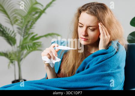 Ein krankes Mädchen mit einer Erkältung oder Grippe in eine blaue Decke gewickelt misst ihre Temperatur zu Hause, Symptome, Behandlung von Viruserkrankungen Stockfoto