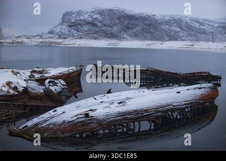Ruinierte Boote ruhen in Teriberka, Russland, halb untergetaucht im Wasser mit Schnee auf ihnen und Bergen im Hintergrund während der frühen Morgenstunden. Stockfoto