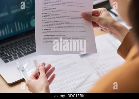 Junge Erwachsene Frau, die Vertragsdokumente hält, während sie Papiere am Schreibtisch überprüft, Laptop mit Datendiagrammen im Hintergrund, Hände und Teilprofil im Fokus Stockfoto