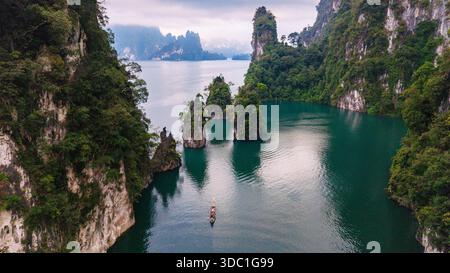 Erkunden Sie das ruhige Wasser des Cheow Lan Lake in Khao Sok, Thailand, umgeben von hohen Kalksteinklippen und üppigem Grün. Ein einsames Boot gleitet friedlich über den ruhigen See und lädt zu Abenteuern ein. Stockfoto