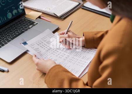 Junge Erwachsene kaukasische Frau, die Finanzdokumente überprüft und Notizen am Schreibtisch schreibt, gedruckte Tabellenkalkulationen in der Nähe eines offenen Laptops hält, Diagramme und Diagramme anzeigt, arbeitet im Büro Stockfoto