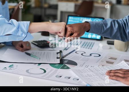 Weiße Frau und Schwarzer Mann, die die Faust über den Schreibtisch werfen, der mit Finanzdiagrammen und einem digitalen Tablet bedeckt ist, zeigen Teamarbeit und Zusammenarbeit in einer modernen Büroumgebung Stockfoto