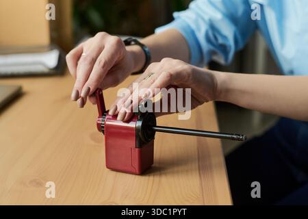 Junge Erwachsene Frau, die Bleistift mit einem manuellen Bleistiftspitzer auf einem Holzschreibtisch schärft, Hände sichtbar, die den Kurbel einstellen, Armbanduhr tragen, natürliche Nägel, Nahaufnahme Stockfoto