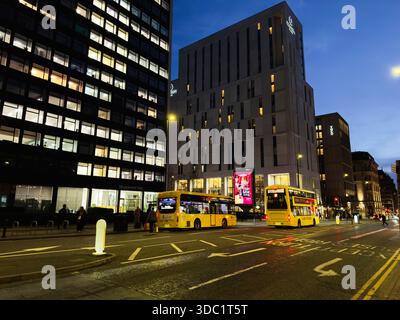Manchester, Großbritannien - 10. Dezember 2025: Geschäftige Nachtszene in Manchester mit gelben Bussen, hohen Büros und beleuchteten Straßen. Stockfoto