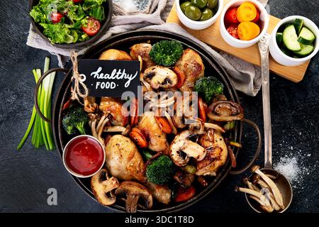 Hähnchenschenkel mit Pilzen, Karotten und Brokkoli in einer Pfanne auf dunklem Hintergrund gebacken. Draufsicht. Stockfoto