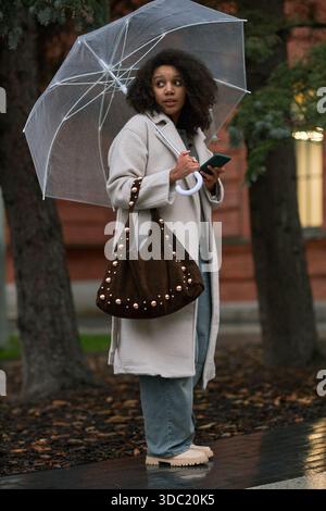 Junge Erwachsene Schwarze Frau, die draußen steht und einen transparenten Schirm und ein Smartphone hält, zur Seite blickt, lange Jacke trägt, große Tasche über der Schulter, Herbstlaub auf dem Boden Stockfoto