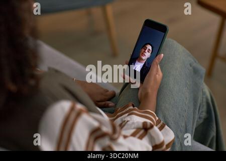 Junger Erwachsener kaukasischer Mann, der während eines Videoanrufs auf dem Smartphone erscheint, junge Erwachsene Frau, die das Telefon hält, während sie auf dem Sofa sitzt, Online-Dating-Kommunikation findet statt Stockfoto