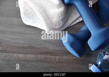 Konzept von Sport, Training und Wellness mit zwei blauen Neopren-Kurzhanteln auf Holzboden mit Wasserflasche und weißem Handtuch. Draufsicht Stockfoto