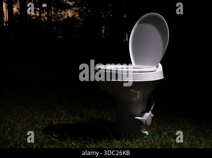 Die Plastiktoilette für Reiseanhänger wurde ausgetauscht und mit offenem Deckel auf einem Feld gelassen, während die Sonne hinter einer dicken Baumlinie untergeht. Stockfoto
