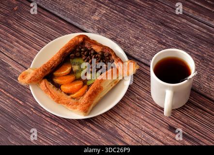 Frisch gebackenes Blätterteigbrötchen mit Kiwi- und Mandarinenstücken und dekoriert mit Kaffeebohnen auf einem Holztisch, daneben eine Tasse heißer Kaffee. Top VI Stockfoto