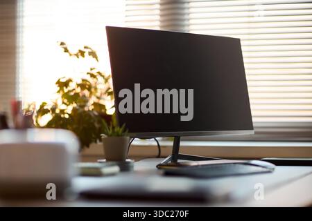 Moderner Computermonitor, der auf einem Schreibtisch neben Tastatur und Maus steht, mit Topfpflanzen und verschwommenen Fensterjalousien im Hintergrund, Technologie-Arbeitsplatzkonzept Stockfoto