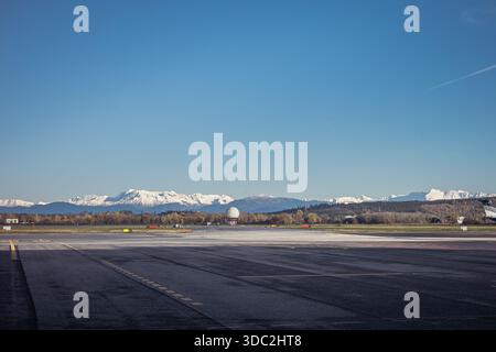 Weite Sicht auf die Start- und Landebahn des Flughafens Triest unter klarem blauem Himmel, mit asphaltierten Rollbahnen im Vordergrund, einer Radarkuppel und Herbstbäumen in der Mitte und Schnee Stockfoto