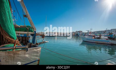 Traditionelle Fischerboote mit verwitterten blauen und weißen Rümpfen säumen das malerische Porto di Trani, während verworrene grüne Netze auf dem Kai ruhen. Der lebendige Stockfoto