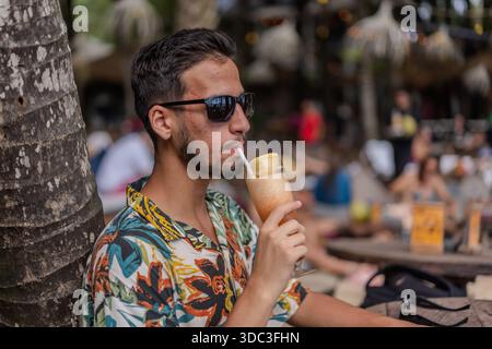 Ein Mann in einem hawaiianischen Hemd trinkt einen Drink mit einem Strohhalm. Er sitzt unter einem Baum Stockfoto