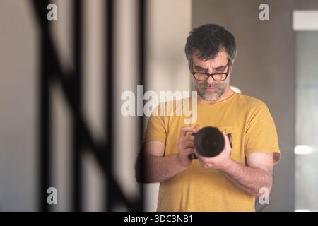 Ein Fotograf trägt eine Brille und ein orangefarbenes T-Shirt, das eine professionelle Kamera hält und ein Selbstporträt vor dem Spiegel in seinem Wohnzimmer macht Stockfoto