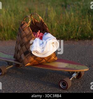 Eine gewebte Strandtasche mit Essentials für den Sommer auf dem Skateboard. Handtuch in einem Korbbeutel Stockfoto