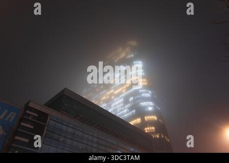 Warmes elektrisches Licht aus Bürokabinen im Kontrast zum kalten, dunklen Nebel, der Einen Glashochhaus umgibt. Bürolichter verwischen sich in Ein leuchtendes Muster Stockfoto