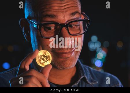 Ein Mann, der nachts eine glänzende Bitcoin-Münze in der Hand sieht, mit Bokeh-Lichtern Stockfoto