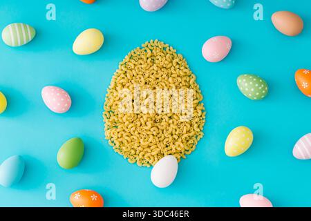 Buntes Muster aus verschiedenen farbigen Ostereiern mit Ostereierform aus rohen Ellenbogen Makkaroni Nudeln auf hellblauem Hintergrund. Stockfoto