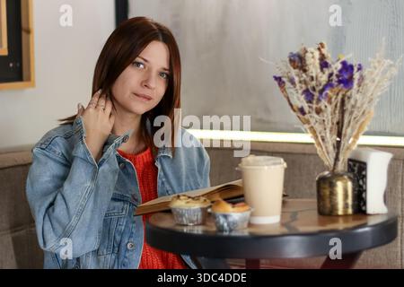 Brünette mit orangefarbenem Oberteil und Jeansjacke sitzt im Café und berührt den Hals, während sie ein Buch liest und Kaffee und Muffins genießt Stockfoto