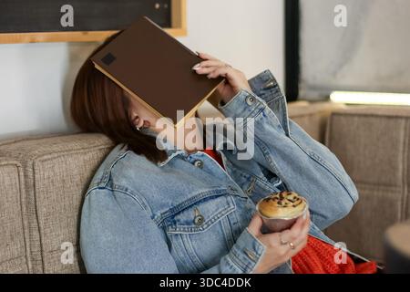 Junge Frau, die das Gesicht mit einem Buch bedeckt, während sie sich zurücklehnt und Schokoladenmuffin im Café hält Stockfoto