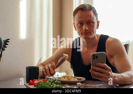 Muskulöser Mann im Tanktop hält Telefon, gabelt Nahrung aus nahrhaftem Gericht mit Gemüse und Nahrungsergänzungsmitteln in der Nähe Stockfoto