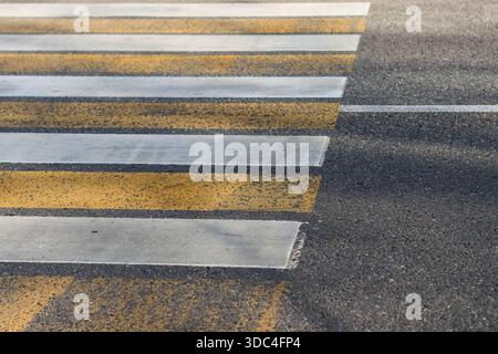 Verwitterter Fußgängerübergang mit abwechselnden gelben und weißen Bändern auf dunkler Asphaltstraße Stockfoto