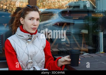 Eine junge Frau in rotem Pullover und grauer Weste sitzt am Kaffeetisch und hält eine schwarze Tasse zum Mitnehmen. Stockfoto