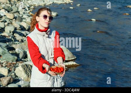 Eine Frau in Sonnenbrille und Weste hält sanft eine kleine Schachtel mit roten Kirschtomaten neben einem klaren Bergbach. Stockfoto