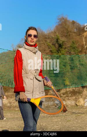Frau in rotem Pullover und beiger Weste hält selbstbewusst orangen Schläger auf einem Schotterplatz mit grünem Netz und hügeliger Kulisse. Stockfoto