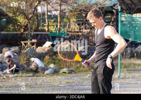 Der muskulöse Mann greift bunte Schläger, steht auf Kies und spielt in der Ferne in der Nähe von Felsen und Netzen. Stockfoto